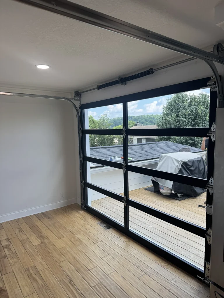 An opened glass garage door revealing an outdoor wooden deck with a covered grill and lush green trees.